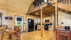 A spacious cabin interior with a wooden dining area, kitchen, and a loft bedroom with a large window.