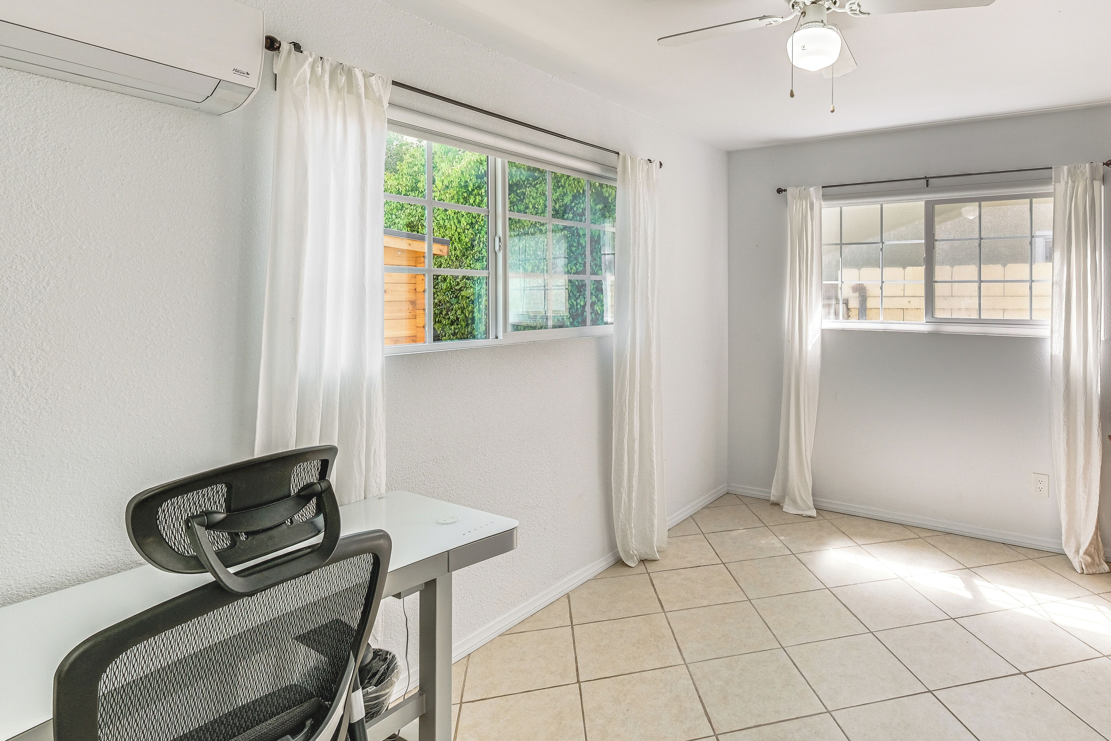 A room with a desk, chair, two windows with curtains, a ceiling fan, and tiled flooring.