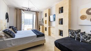 A modern hotel room with a large bed, a smaller sofa, a TV mounted on a stone wall, and a view of the outdoors. - Cala Coralli (Syracuse)