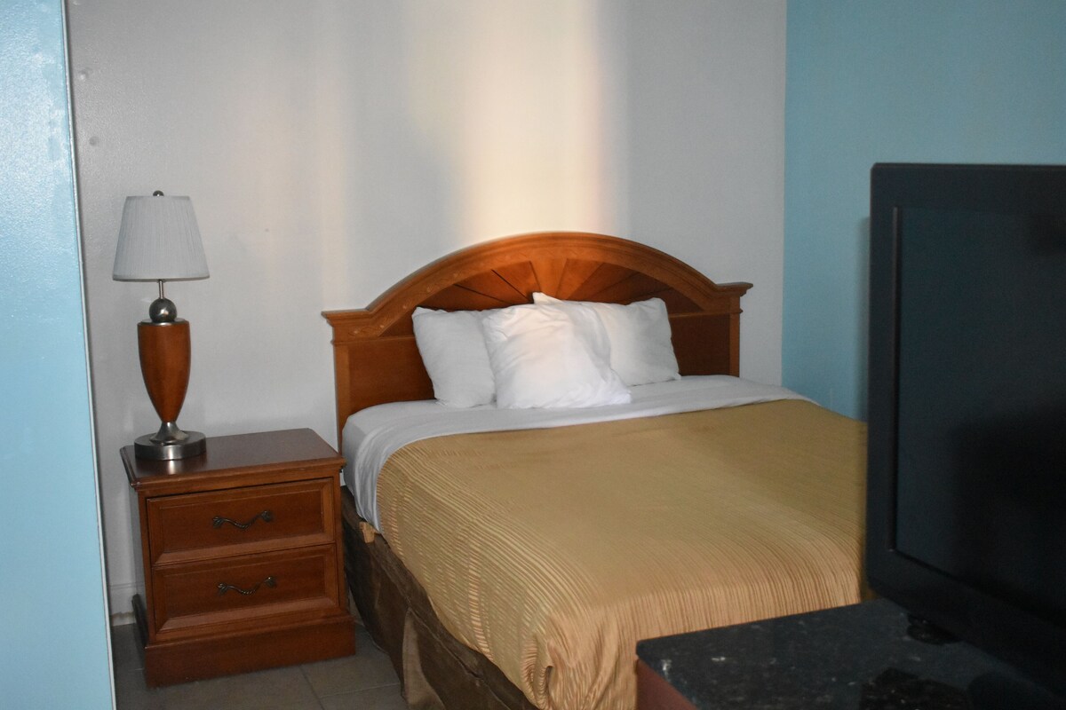 A hotel room with a wooden bed, a nightstand with drawers, a lamp, and a television.