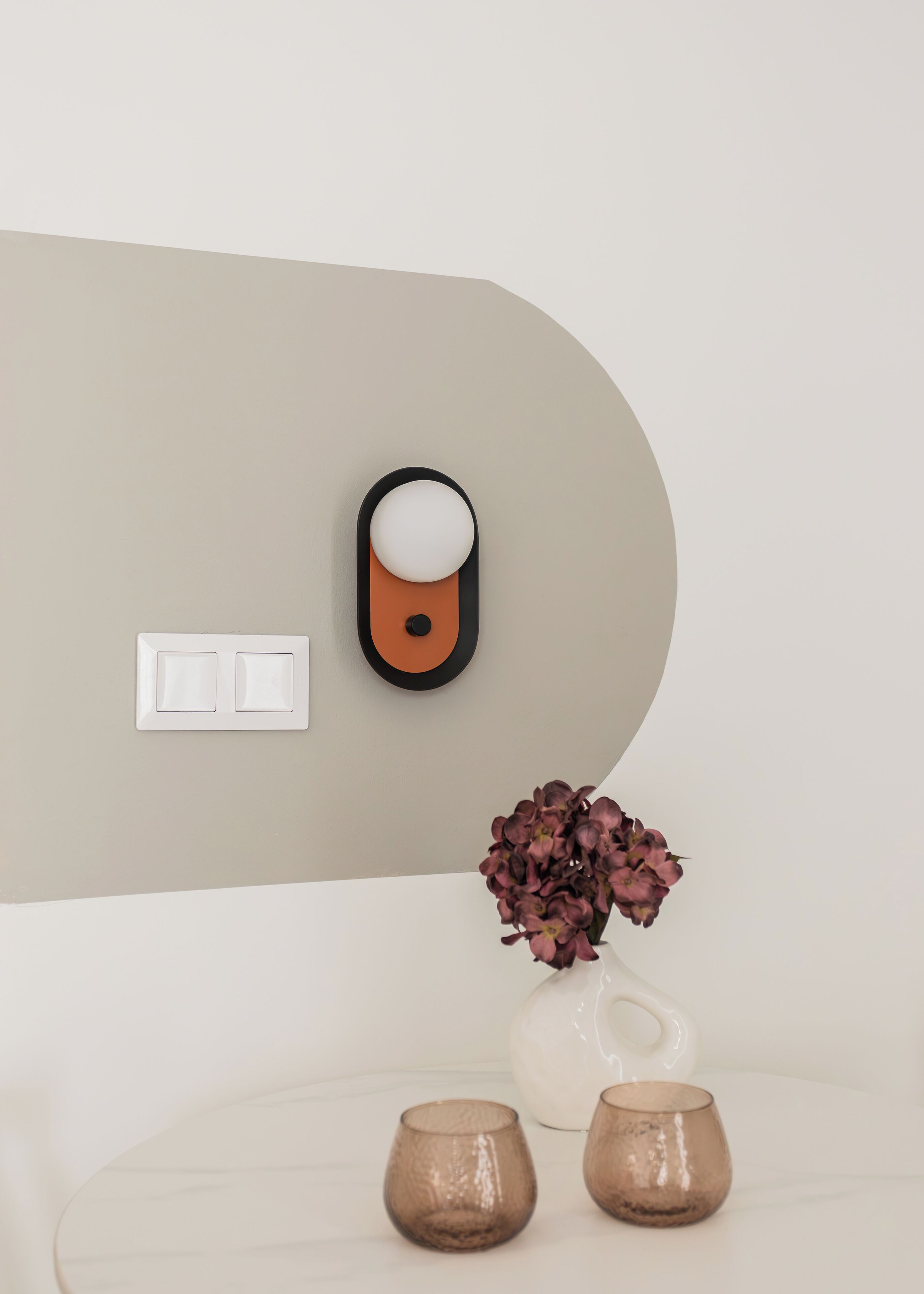 A minimalist room with a wall-mounted light fixture, a vase with flowers, and two small cups on a table.
