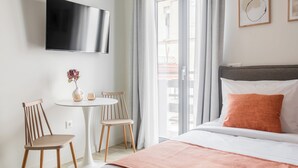 A hotel room with a bed, a TV mounted on the wall, a small round table with chairs, and a view of the outside through a sliding door. - SOL by Zoia Living (Athens)