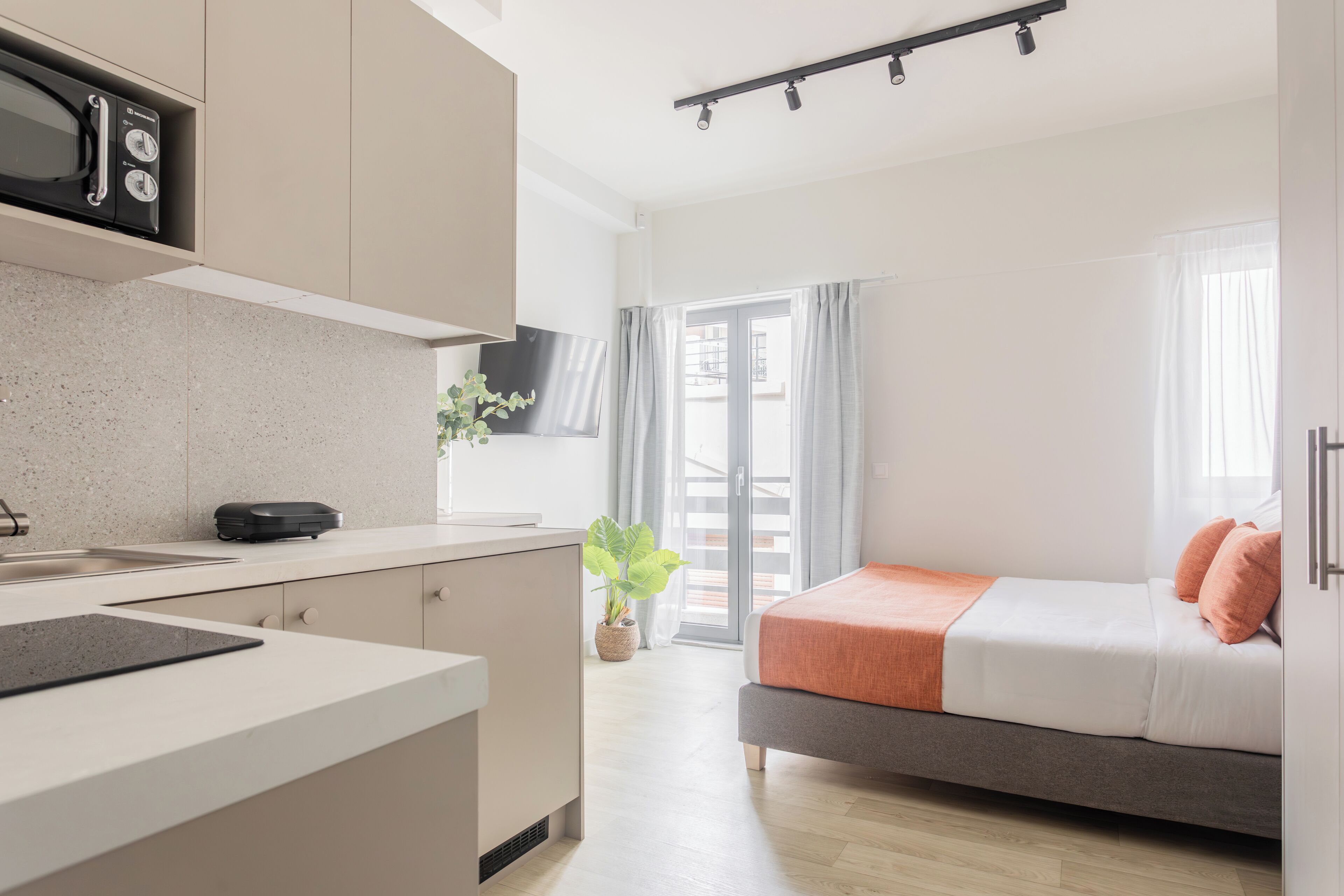 A modern kitchen with a microwave, a sink, and a countertop. There is a bed with a gray and white mattress and orange pillows. A plant is on the windowsill, and a sliding glass door leads to an outdoor area.