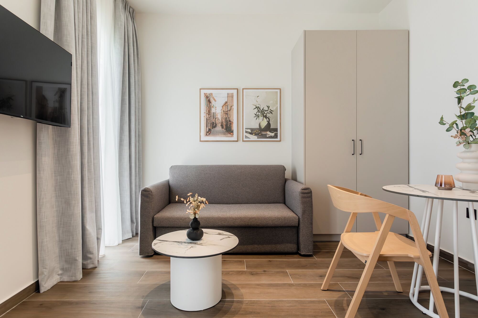A modern living room with a grey sofa, a round coffee table with a vase, a wooden chair, a TV mounted on the wall, and two framed pictures.