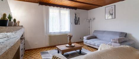 Une pièce chaleureuse avec un plafond en bois, un canapé gris, un lit blanc et une petite table sur laquelle est posé un vase.