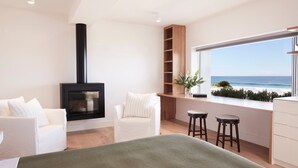 Un salon moderne avec un poêle à bois noir, un canapé blanc, un plaid vert et une vue sur l’océan par une grande fenêtre.