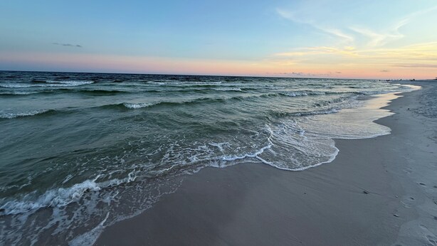 A beach with waves gently lapping onto the shore, a clear sky, and distant buildings.