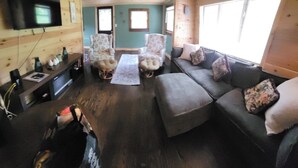 A cozy cabin interior with a wooden ceiling, a TV mounted on the wall, a sofa, two patterned armchairs, and a coffee table.