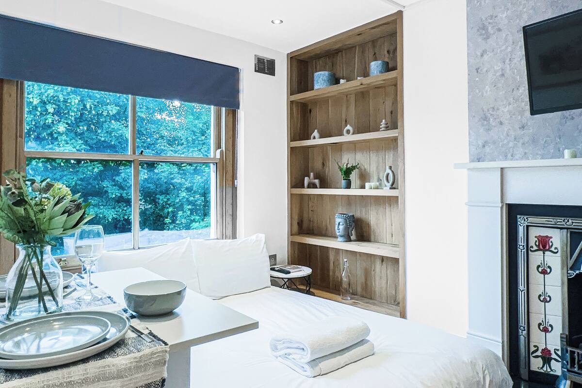 A modern bedroom with a large bed, a wooden bookshelf, a fireplace, and a window with a view of trees.