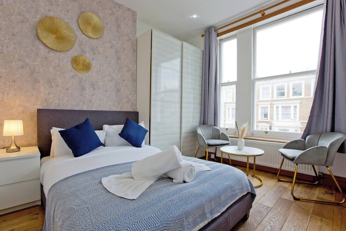 A hotel room with a large bed, a small table, two chairs, and a view of buildings through the window.
