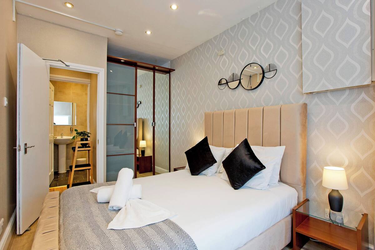 A hotel room with a large bed, a bedside table with a lamp, a mirror on the wall, and a bathroom visible through an open door.