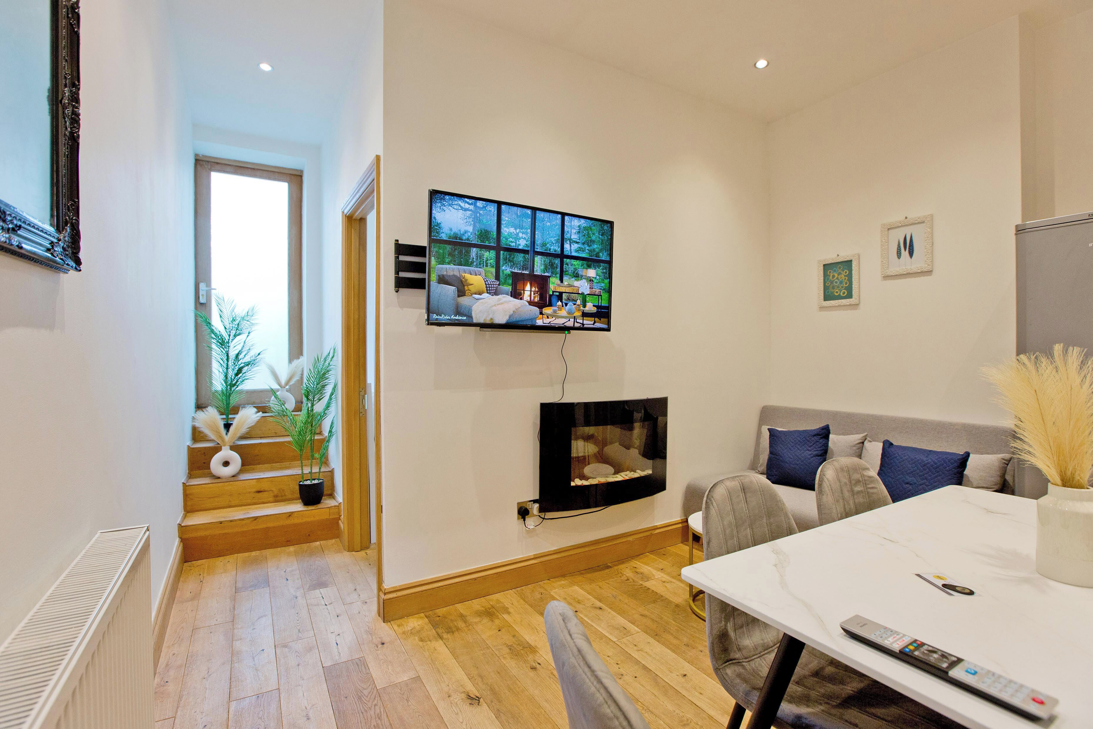 Una sala moderna con televisor de pantalla plana montado en la pared, un sofá con cojines azules, una mesa de comedor con un control remoto y una escalera adornada con plantas en macetas.