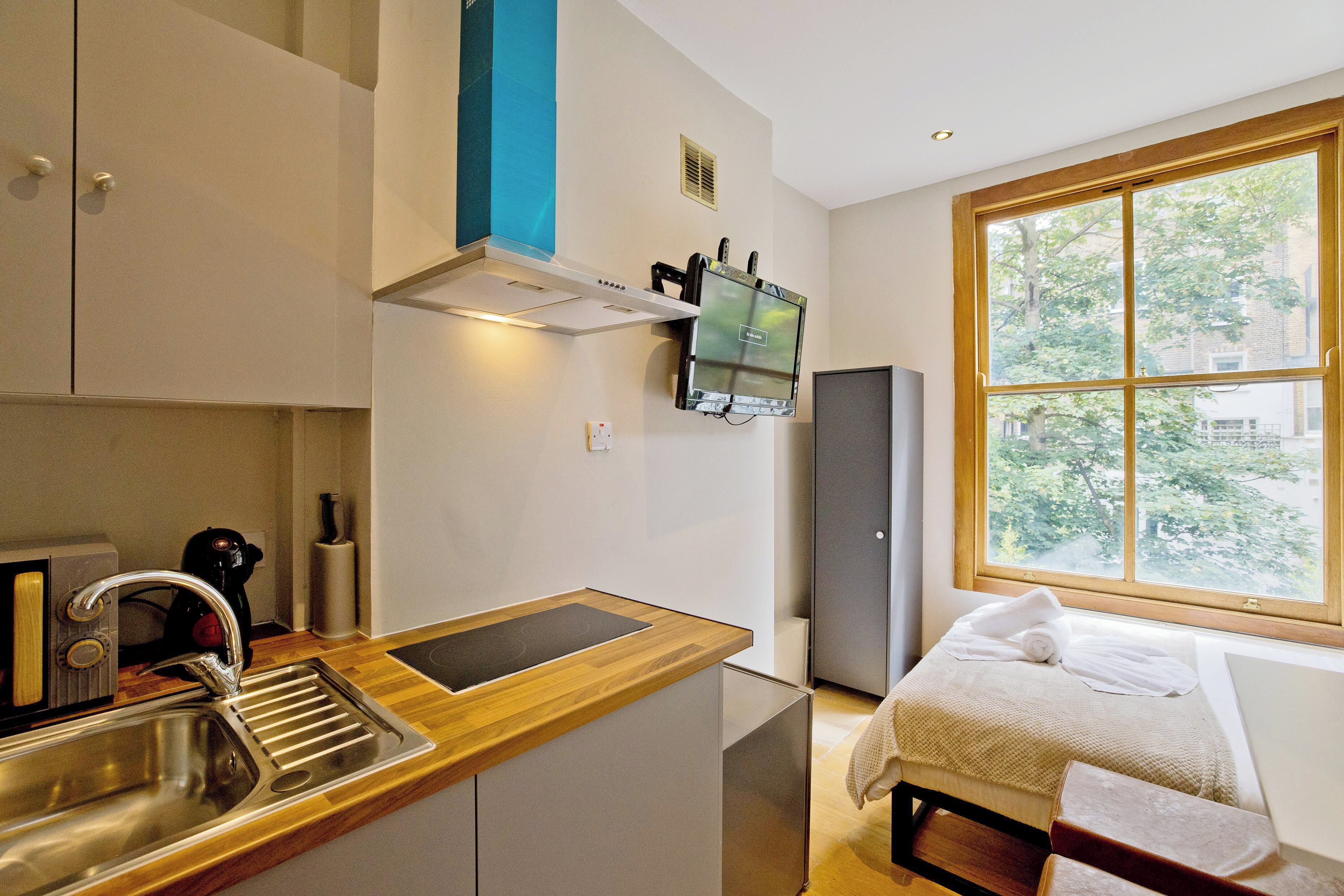 A compact kitchen with a wooden countertop, stainless steel sink, and a built-in range hood. A small refrigerator is visible in the background, and a window provides a view of the outdoors.