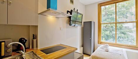 A compact kitchen with a wooden countertop, stainless steel sink, and a built-in range hood. A small refrigerator is visible in the background, and a window provides a view of the outdoors.