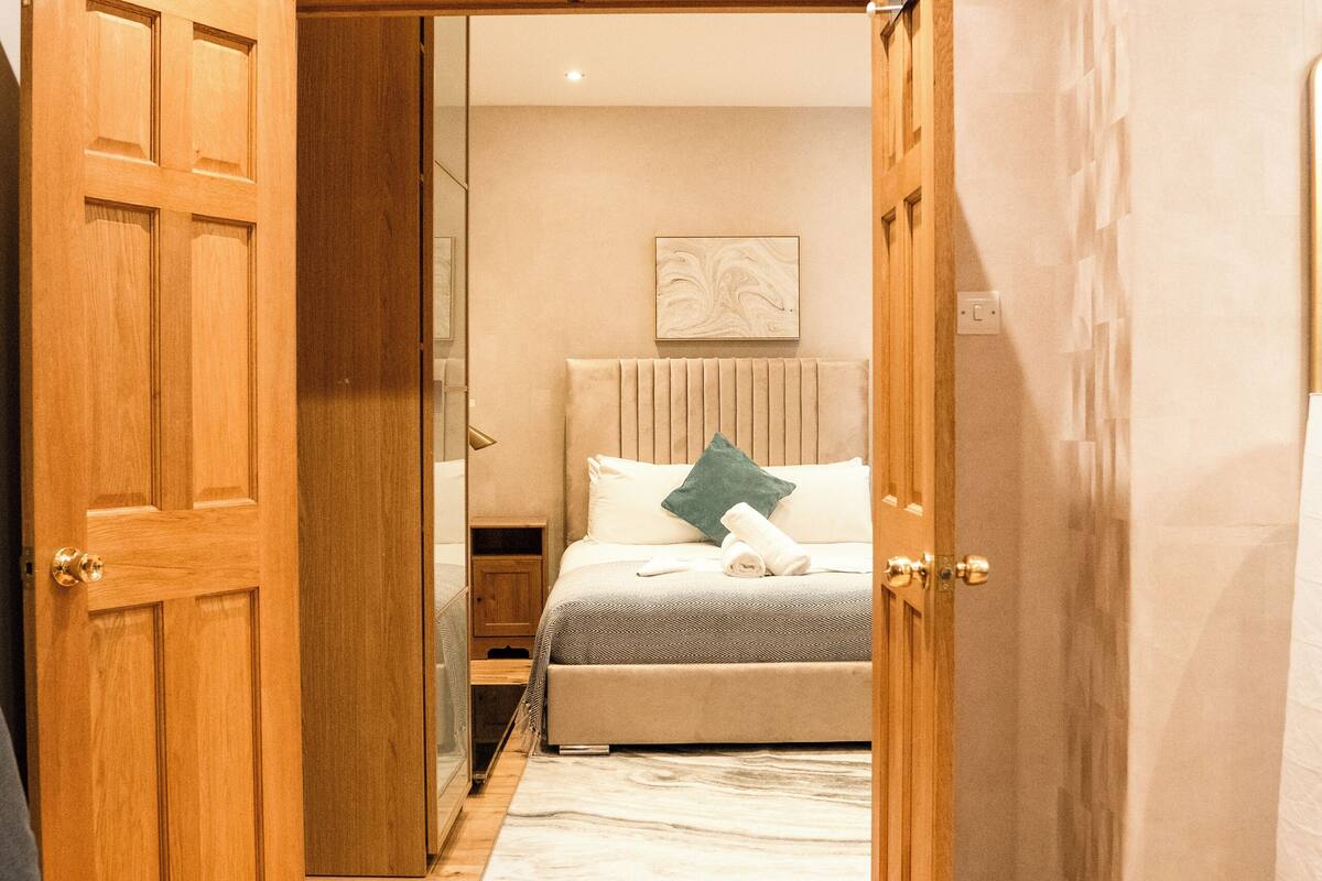 A hotel room with a wooden door, a bed with a white headboard, a gray sofa, and a wooden floor.