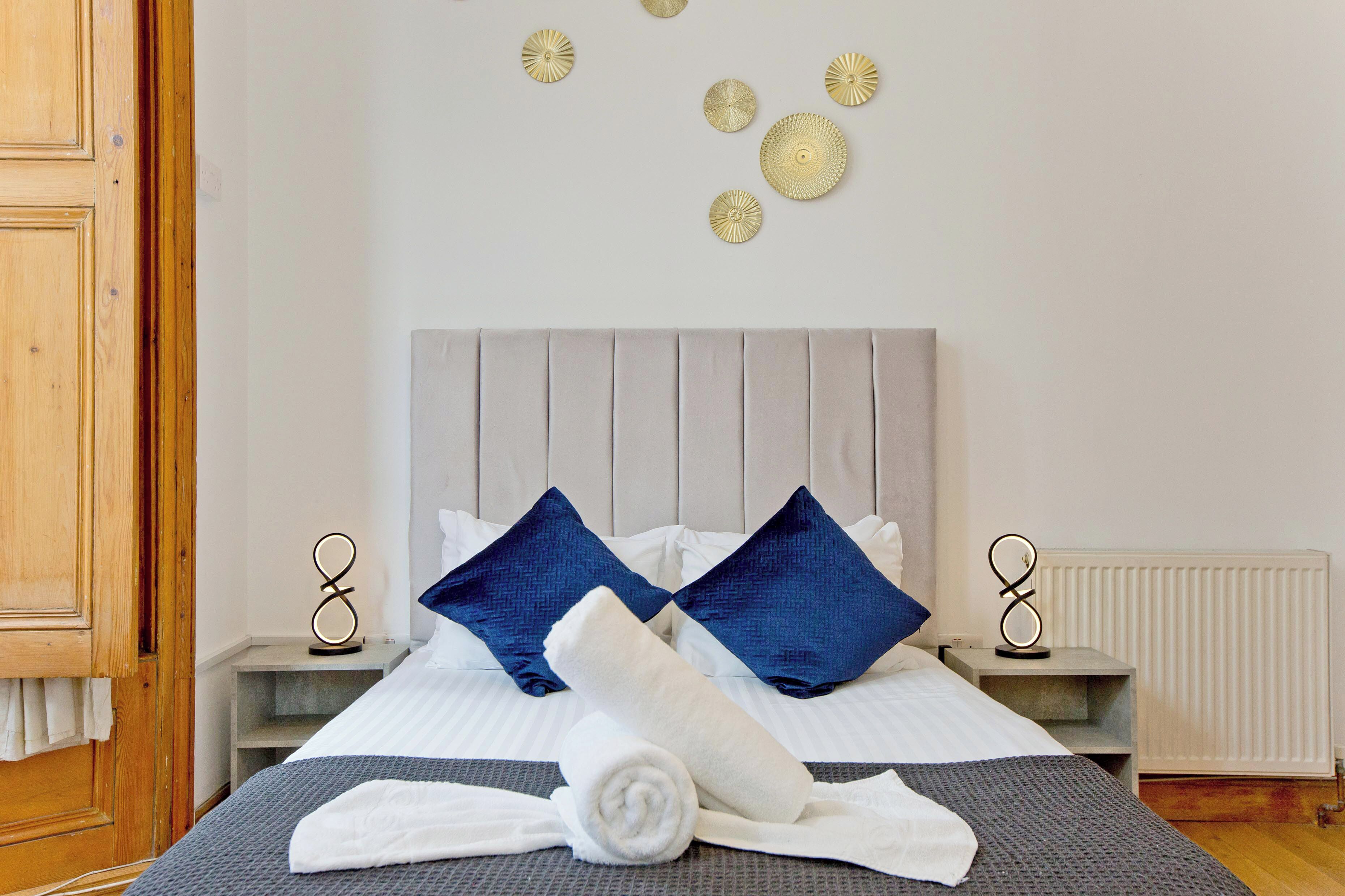 Uma cama bem arrumada com almofadas azuis e brancas, uma toalha branca enrolada e arte decorativa na parede.
