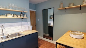 Una cocina con un lavamanos doble, armarios oscuros, una mesa de madera y estantes con cristalería y utensilios de cocina.