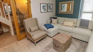 Una sala de estar con un sillón color beige, un sofá de tonos claros, un reposapiés de mimbre y un tapete estampado. Hay cuadros enmarcados en la pared y un espejo sobre el sofá.