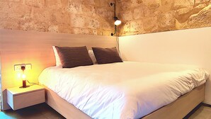 Un dormitorio con cama, dos almohadas, una mesita de noche con lámpara y una pared de piedra.