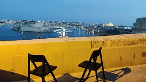 Dos sillas negras frente a una pared amarilla, con vista a un puerto con barcos y edificios.