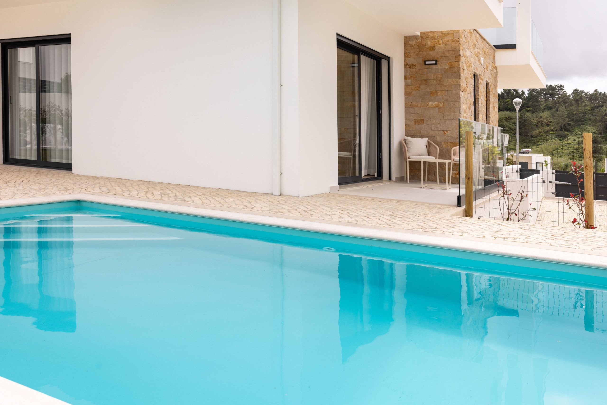 A modern poolside area with a clear blue pool, a stone wall, and a seating area with a chair and table.