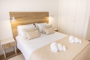 A neatly made bed with white linens and beige pillows, a bedside table with a lamp, and a set of rolled towels on the bed. - Encanto da Foz - BY SCH (Foz do Arelho)