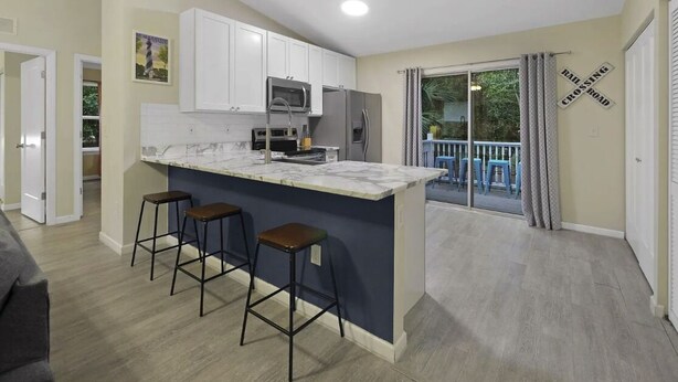 A modern kitchen with white cabinets, a marble countertop, and a built-in oven. There are two bar stools and a large window with a view of a balcony. - Charming 3BR in St. Augustine - Walk to Downtown, Near Beach, Wifi & A/C! (St. Augustine)