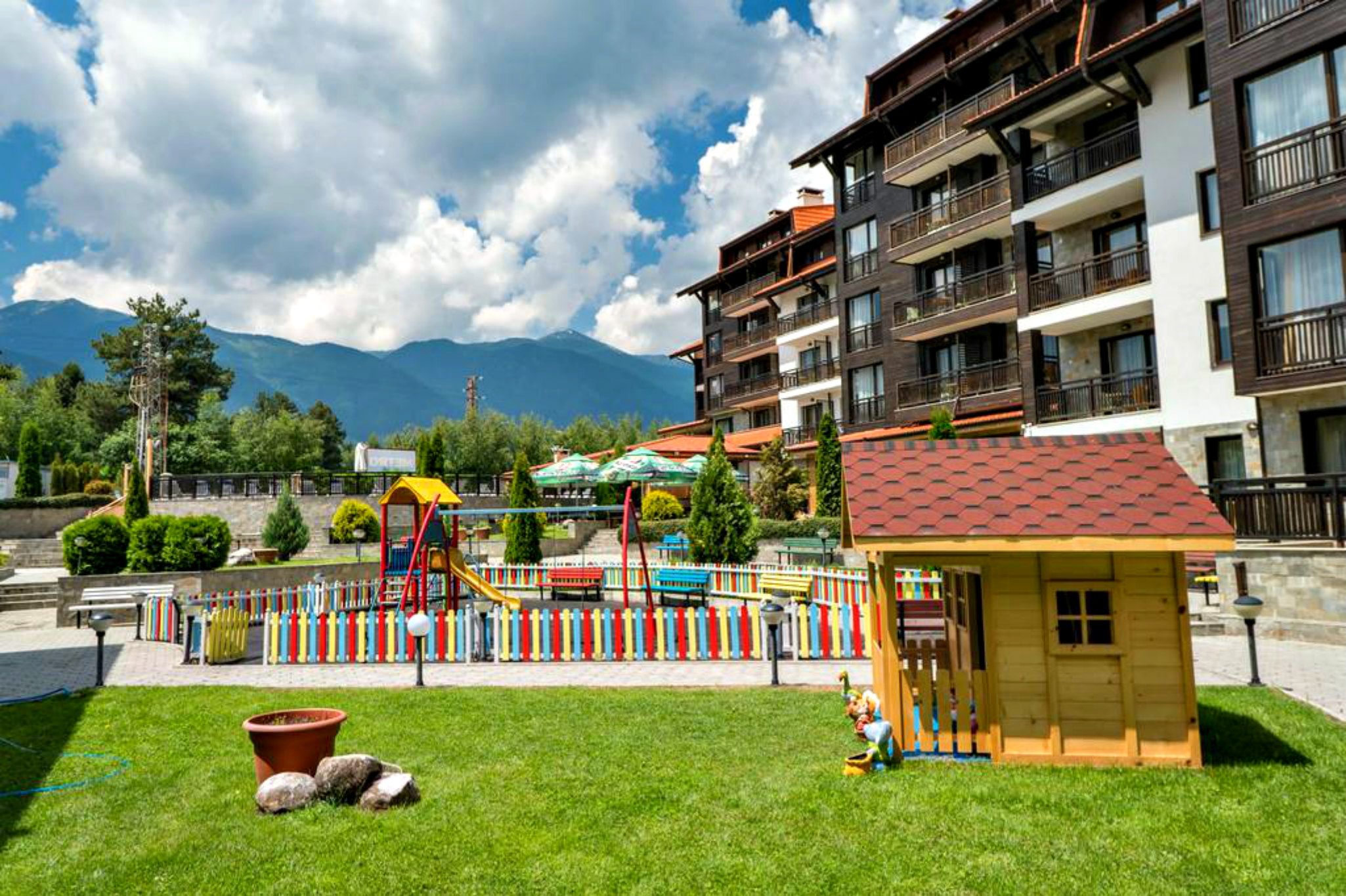 A playground with slides and climbing structures, a small wooden playhouse, and a building with balconies and a red roof.
