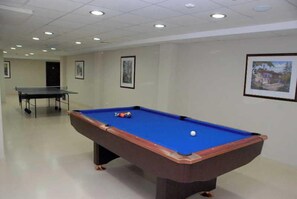 Une table de billard avec des billes, une table de ping-pong et des tableaux encadrés accrochés aux murs, dans une pièce dont le plafond est recouvert de carreaux et les murs sont blancs.