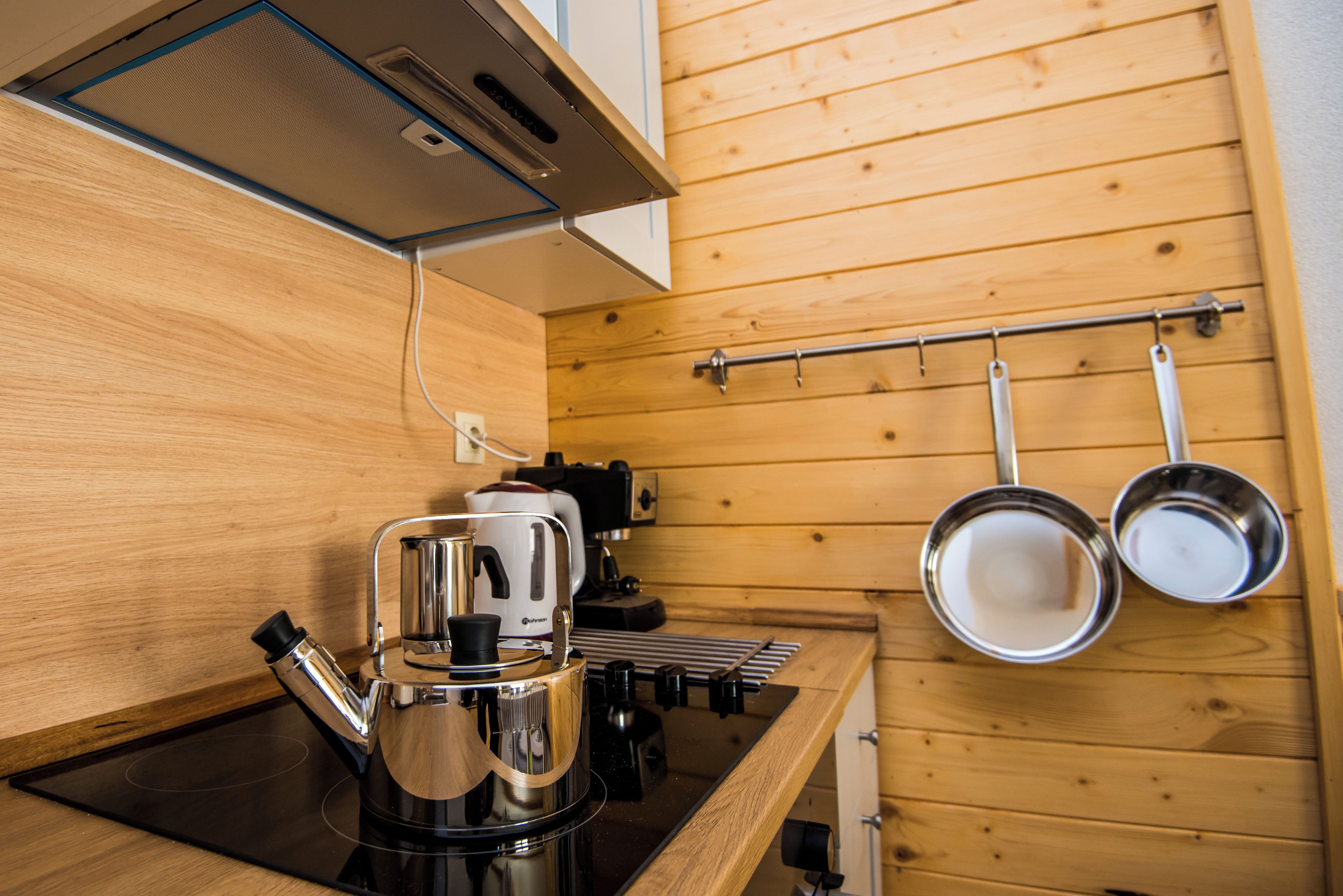 A compact kitchen with a coffee maker, kettle, and hanging pots on a rack.