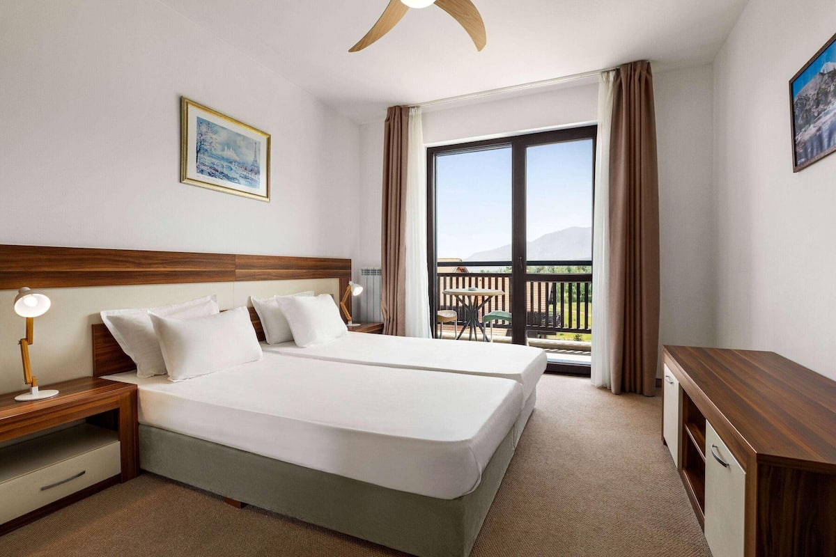 A hotel room with two beds, a desk, a chair, and a balcony with a view.