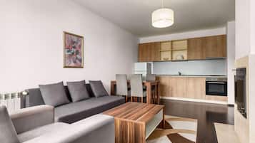 A modern living room with a sofa, a wooden coffee table, and a dining table with chairs. There is a kitchen area with wooden cabinets and a built-in oven.
