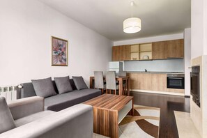 A modern living room with a sofa, a wooden coffee table, and a dining table with chairs. There is a kitchen area with wooden cabinets and a built-in oven. - The Balkan Jewel Resort, Trademark Collection By W (Razlog)