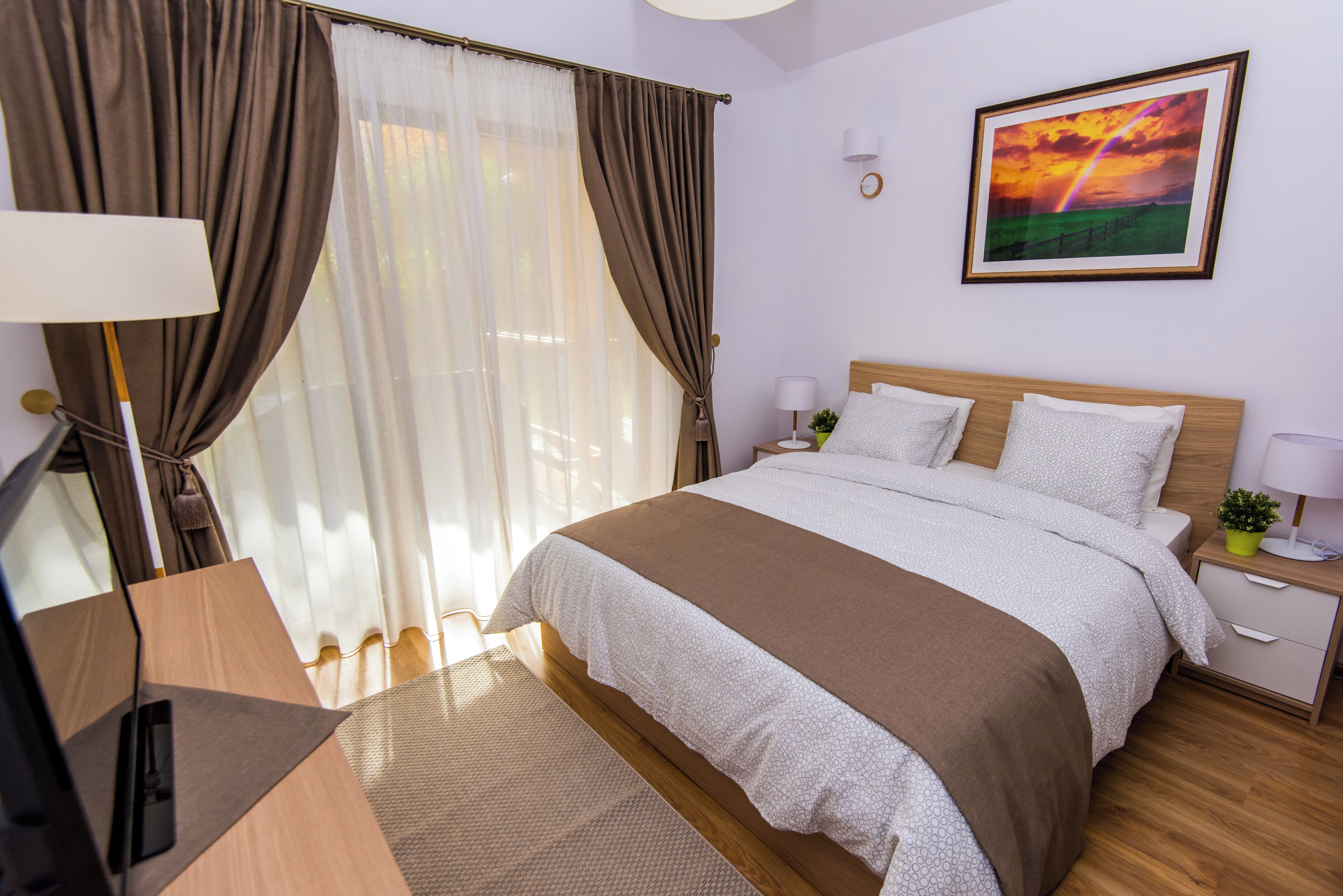 Ein Hotelzimmer mit einem Bett, Vorhängen, einem Schreibtisch, einer Lampe, einem Nachttisch, einer Topfpflanze und einem gerahmten Bild an der Wand.