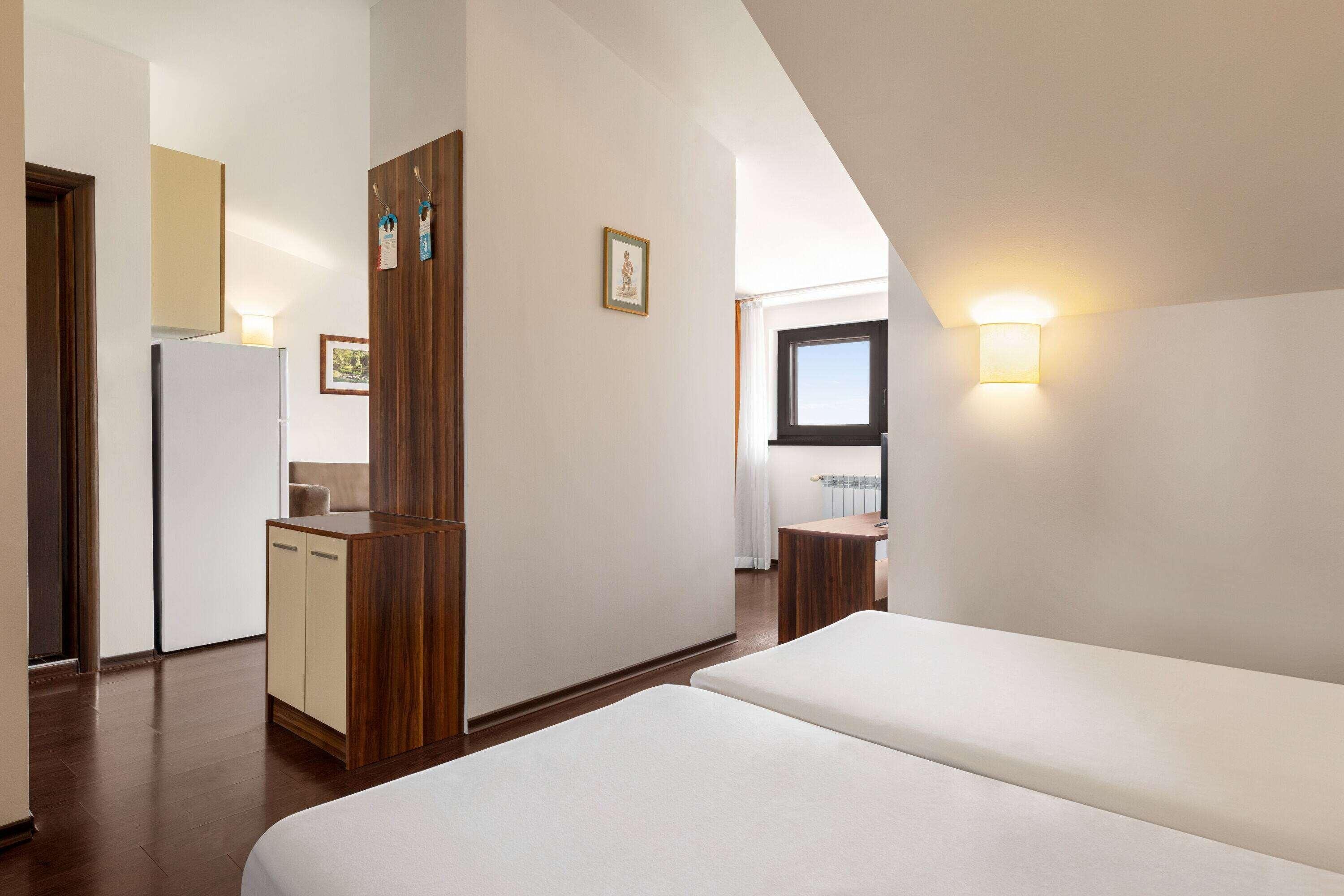 A hotel room with a bed, a wooden wardrobe, a small kitchenette, and a bathroom with a window.