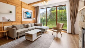 Ein modernes Wohnzimmer mit einem Ecksofa, einem Couchtisch, einem Sessel, einem Kamin und einem großen Fenster mit Blick ins Grüne.