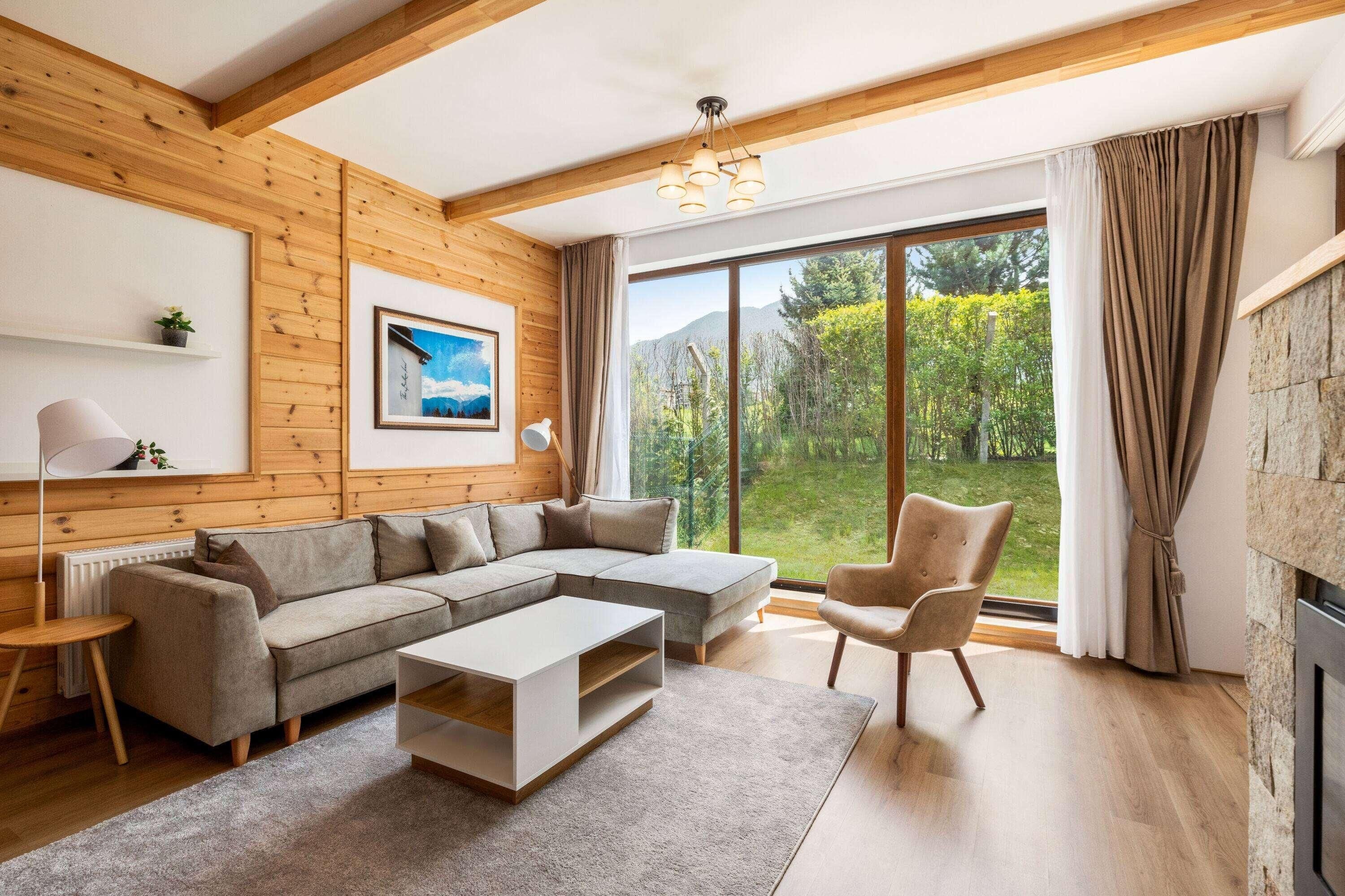 Een moderne woonkamer met een hoekbank, een salontafel, een fauteuil, een open haard en een groot raam met uitzicht op groen.
