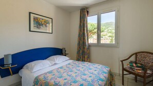 A bedroom with a bed, a chair, a nightstand, a window with a view, and a framed picture on the wall.