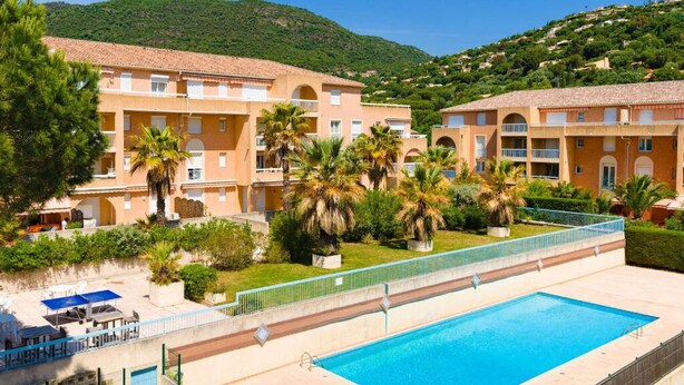 A hotel with a swimming pool, surrounded by palm trees and greenery, with a mountainous backdrop.