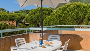A round table with white chairs, a bottle, glasses, and a plate of food on a balcony with a white railing, overlooking a mountainous landscape.