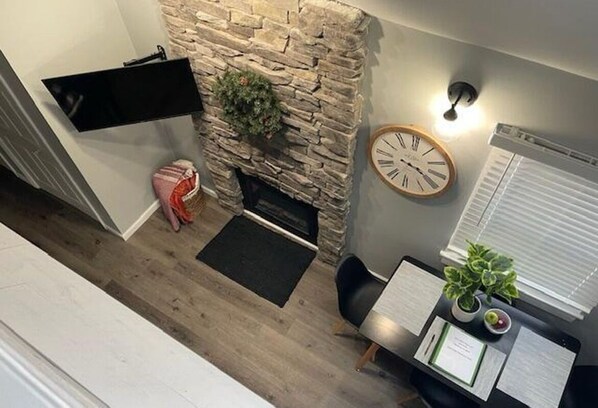 Una habitación con chimenea de piedra, televisor montado, un reloj, un escritorio con computadora y una planta en maceta.