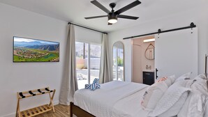 Een slaapkamer met een bed, een plafondventilator, een houten rek, een groot raam met gordijnen en een schilderij van een landschap.