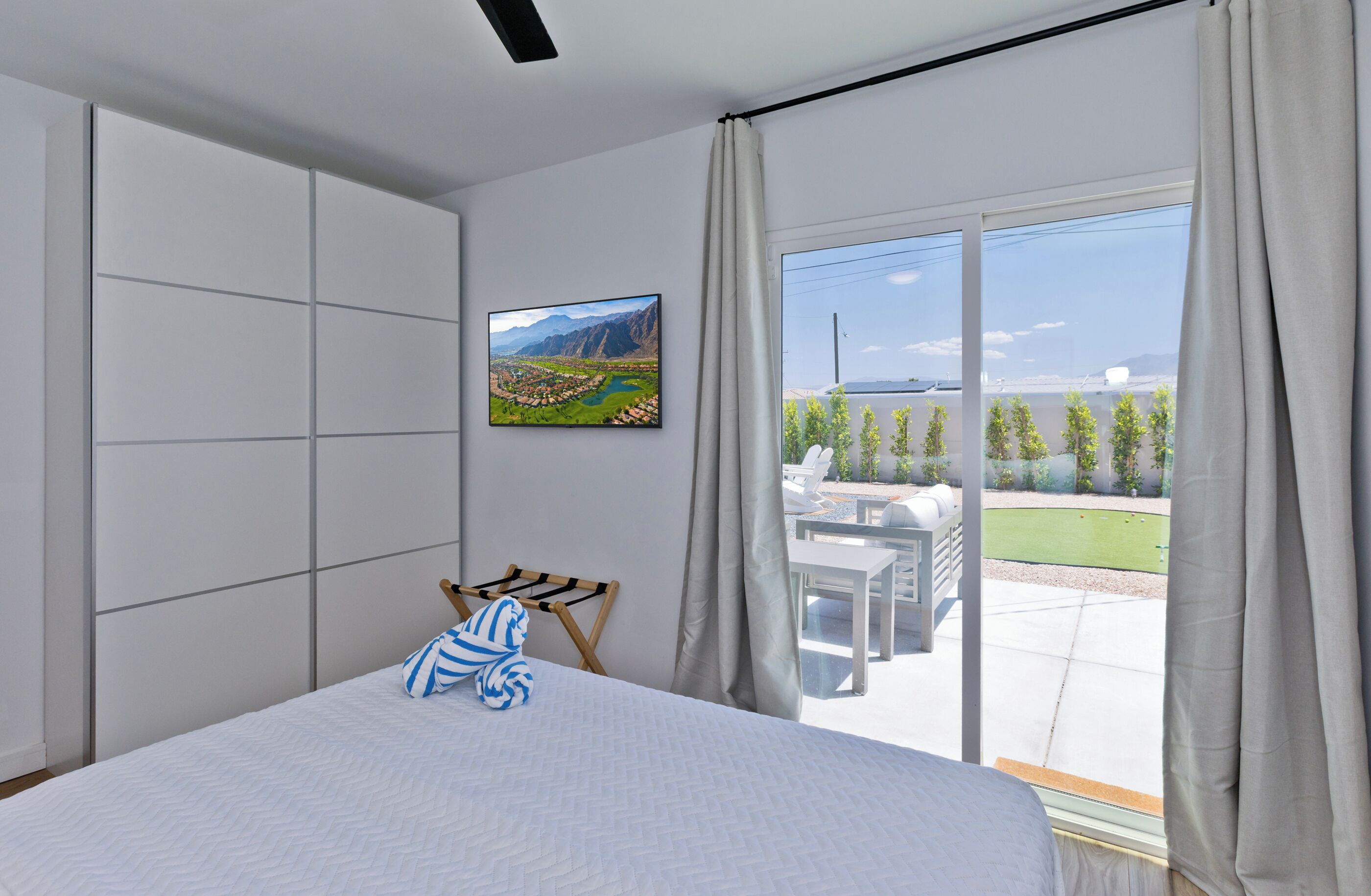 A bedroom with a large bed, a view of a landscaped outdoor area, and a mounted picture on the wall.