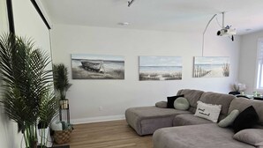 Una sala de estar moderna con un sofá modular gris, dos cuadros colgados en la pared que muestran un bote en el agua, un proyector instalado en el techo y una planta grande en maceta.