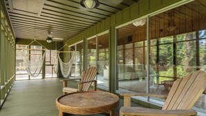 A wooden deck with a round wooden table and two Adirondack chairs, large windows, and a hammock.