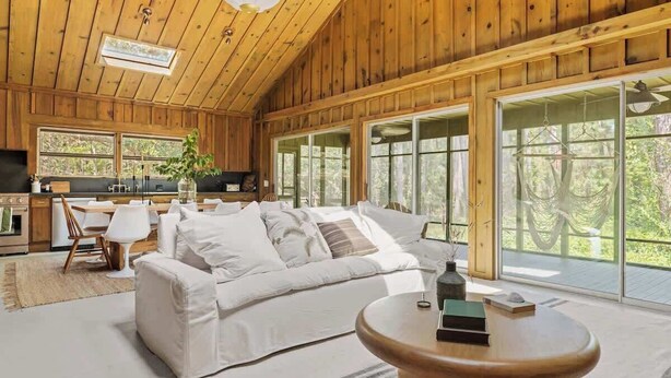 A spacious living area with a white sofa, a wooden coffee table, and large windows offering a view of the outdoors.