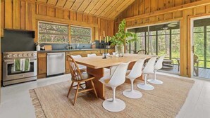 A wooden cabin interior with a large dining table, white chairs, a kitchen with a stove, and a view through large windows.