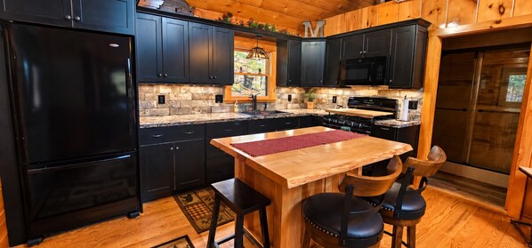 A kitchen with black cabinets, a wooden island, a black refrigerator, and a stone backsplash.