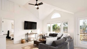 Amplia sala de estar con un sofá gris, un televisor de pantalla plana montado en la pared, un ventilador de techo y ventanas grandes con vistas al exterior.