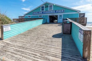 Uma passarela de madeira leva a um edifício turquesa com a placa 'Avalon Fishing Pier'.